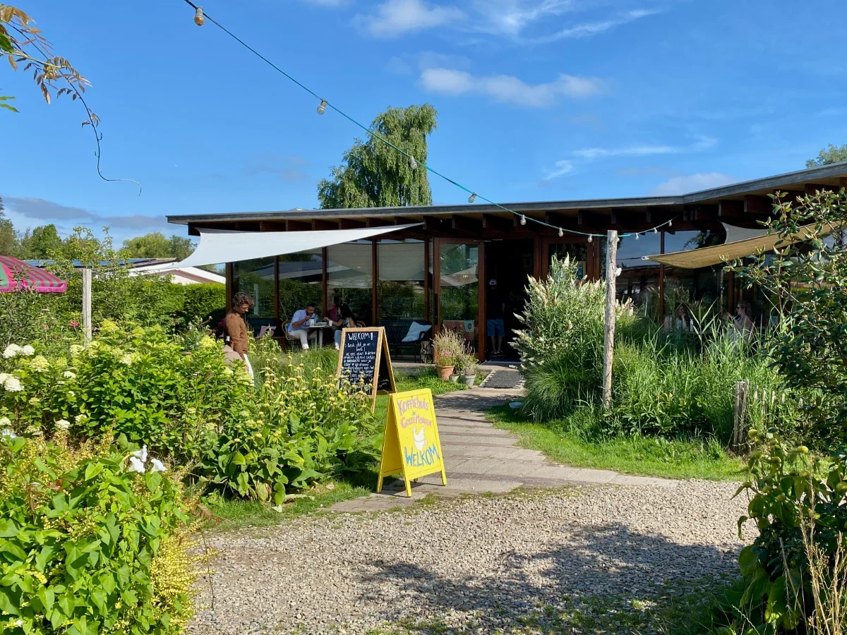 Cafe surrounded by flowers and greenery on a sunny day with string lights, outdoor seating, and sandwich board signs