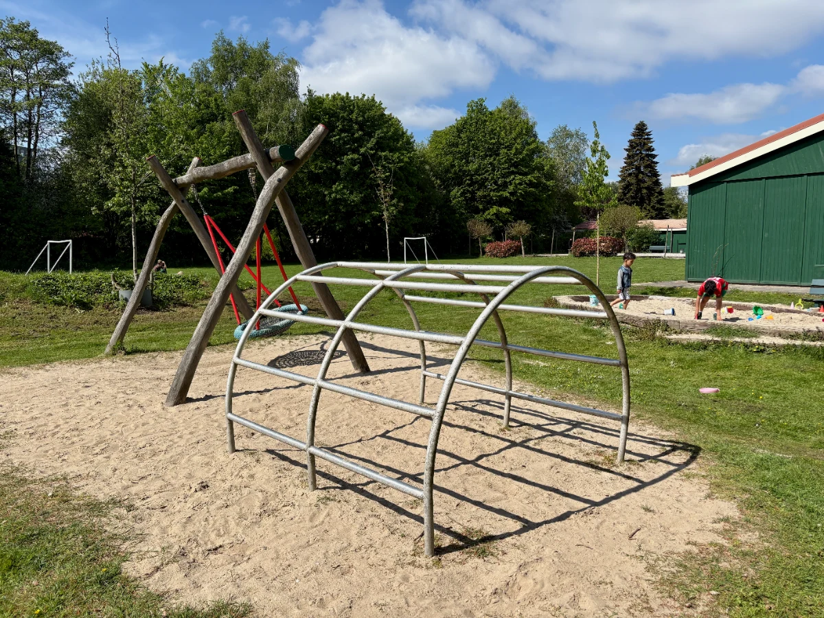 Sunny playground with arched climbing frame, nest swing, and sandbox with green field and trees behind