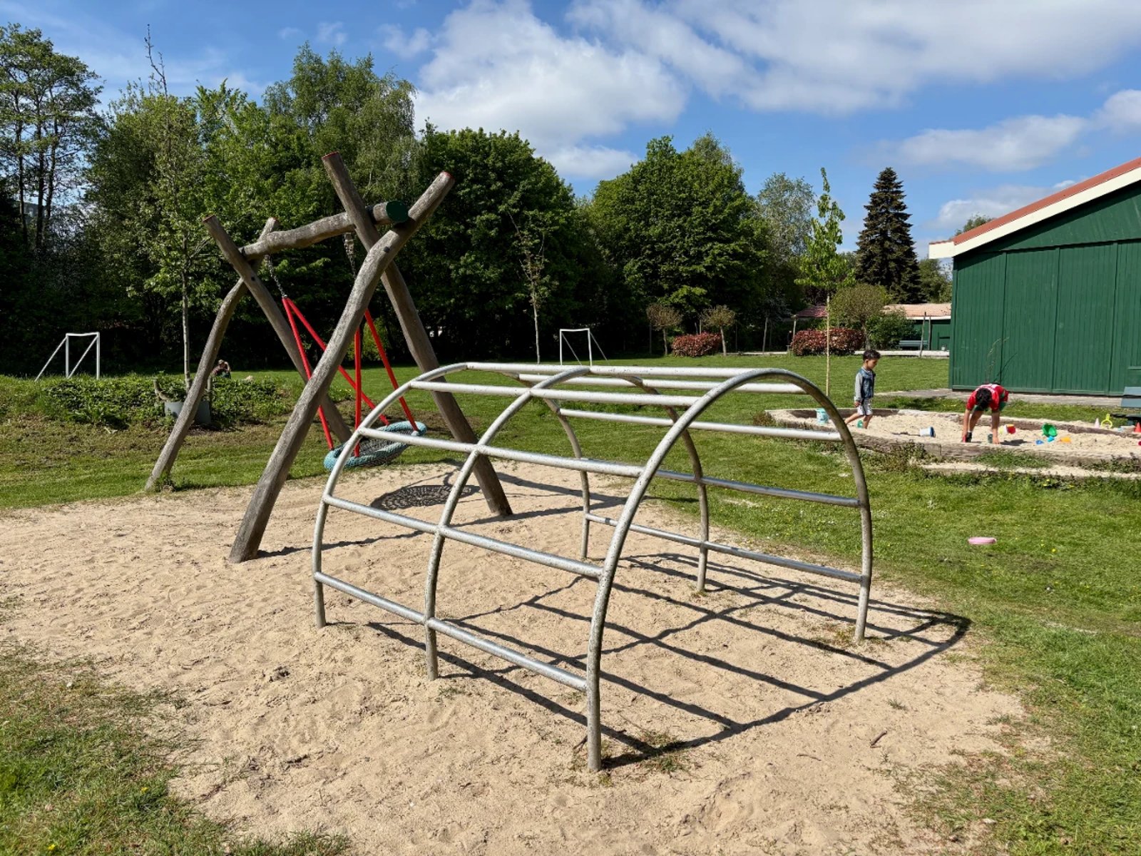 Sunny playground with arched climbing frame, nest swing, and sandbox with green field and trees behind