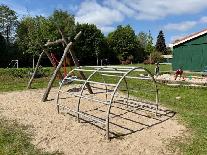 Sunny playground with arched climbing frame, nest swing, and sandbox with green field and trees behind