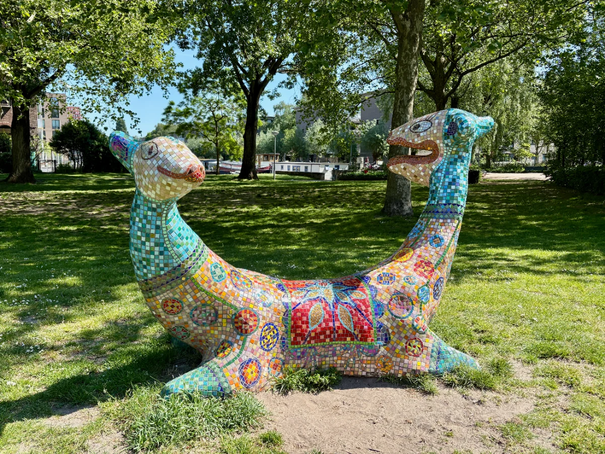 Two-headed animal sculpture covered in colorful mosaic tiles in park setting