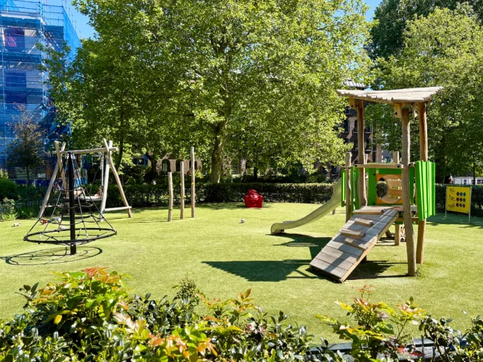 Small, sunny playground with small climbing structure and slide and climbing pyramid and nest swing behind