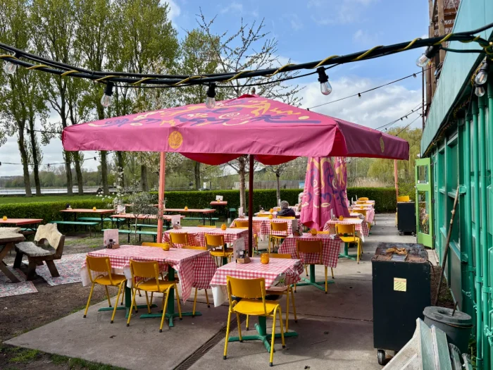 Large pink umbrella shades terrace with red checkered table cloth-covered tables and yellow chairs in a park setting with string lights overhead