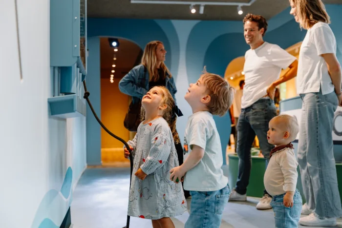 Girl holding rope attached to toy on the wall while other kids look on and parents chat in the background