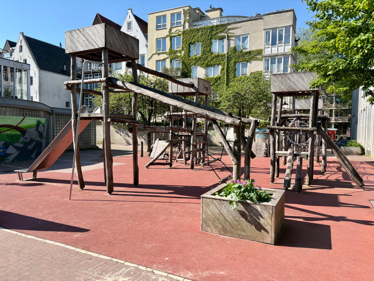 Wooden climbing structure with slide, bridges, and ladders set on red rubberized ground covering and with ivy covered building behind