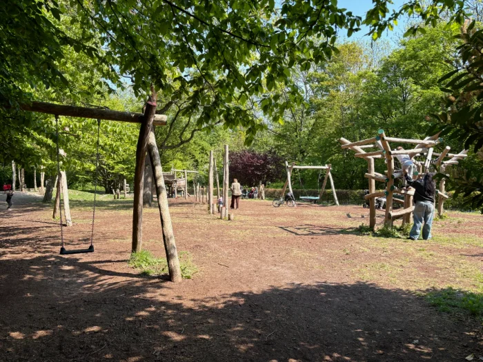 Kids and parents play on shaded playground with swing and natural climbing structures