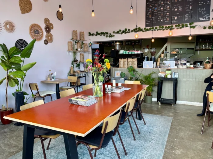 Coffee shop interior with large red work table, flowers, and plants and sage green coffee bar and chalkboard menu above