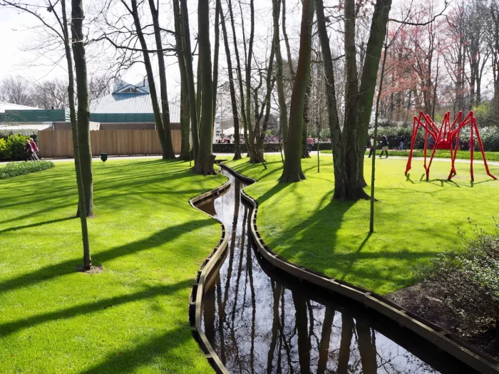 Perfectly manicured lawn with narrow curvy canal, trees, and modern red sculpture of a spider