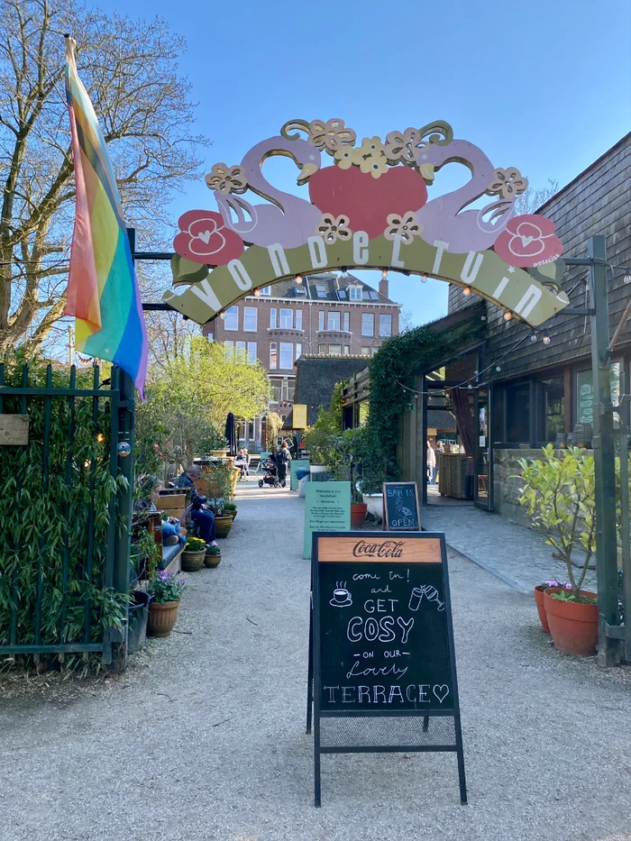 Colorful sign with flowers and swans over entrance gate to De Vondeltuin with outdoor terrace seating and cafe beyond 
