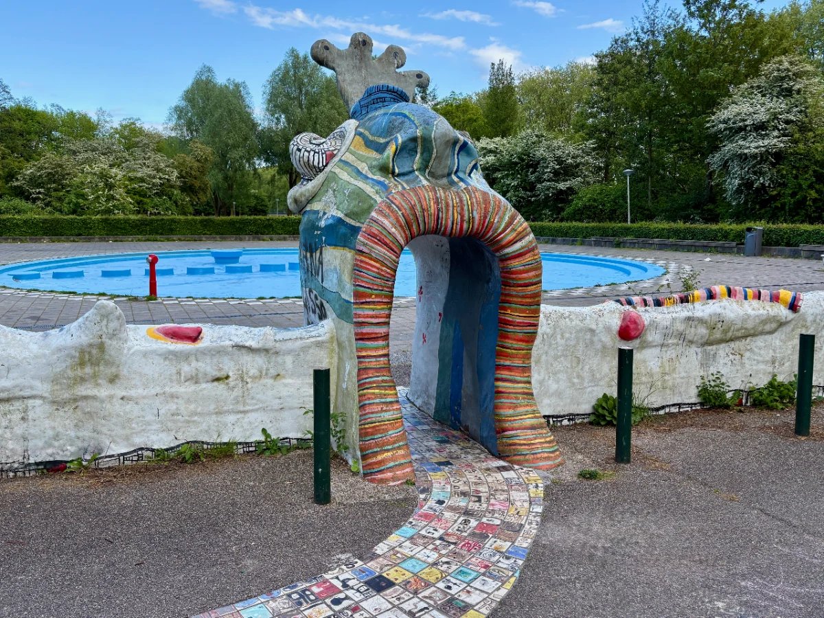 Funky sculptural portal covered in colorful tiles leading to a bright blue paddling pool