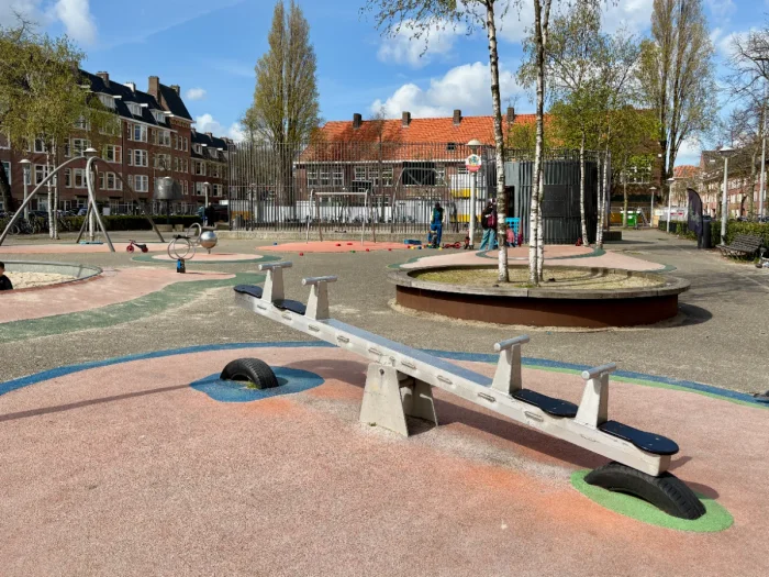 Paved playground on sunny day with seesaw, swings, and fenced sports court behind