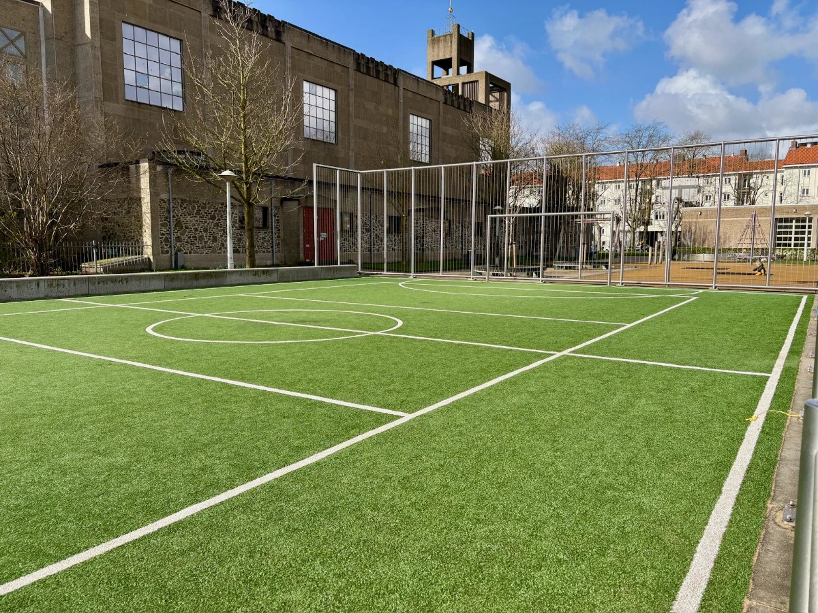 Sunny football pitch with astroturf and white lines surrounded by an enclosure and buildings