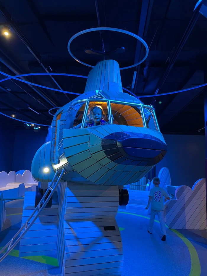 Girl sitting in pretend helicopter in a blue-lit indoor play space 