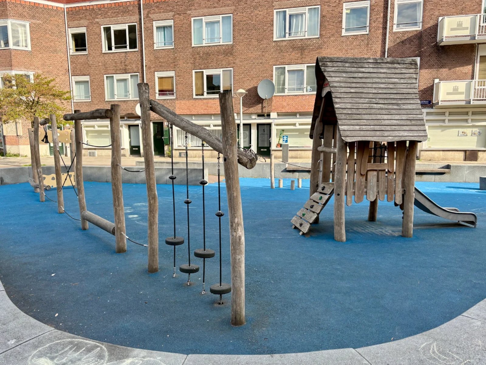 Wooden play house with slide next to wood and rope balance obstacle course set on blue rubber play surface