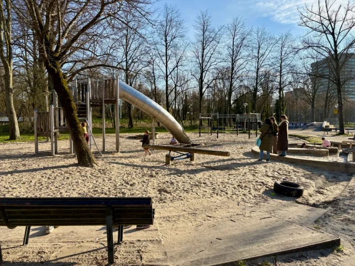 Children and parents at a playground with climbing structure and slide, seesaw, lots of sand and bench under mature trees
