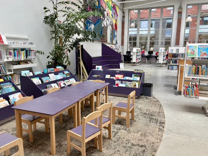 Children's table and chairs, shelves with children's books, and rows of books in a bright, airy library with colorful artwork on the wall