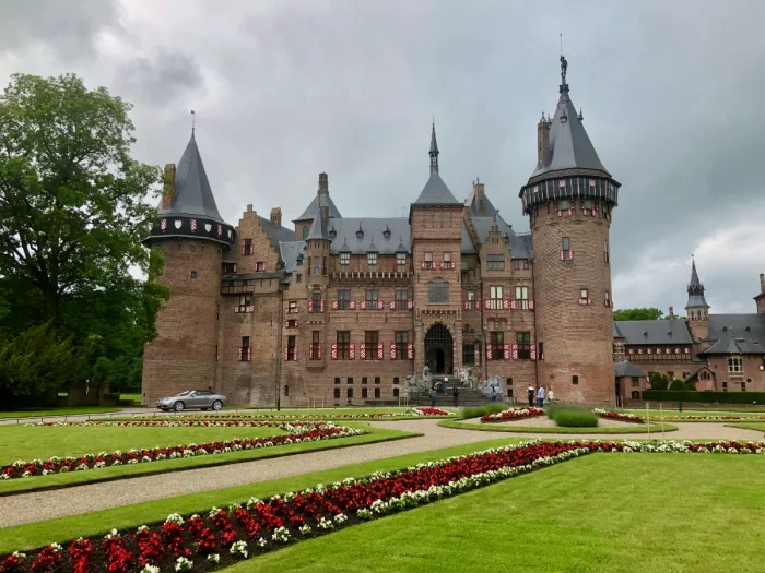 Stately brick castle with grey slate roof, red and white shutters, and manicured garden with red and white flowers