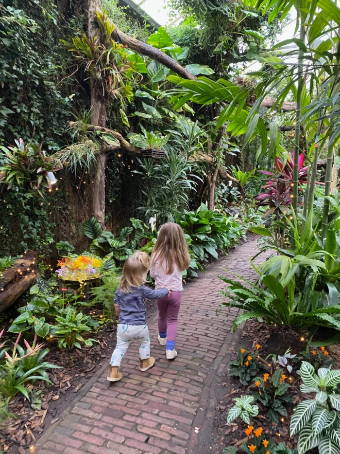 Two children walk along a brick path through a tropical greenhouse with plants and flowers