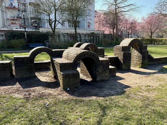 Brick play structure resembling ruins with walls and arches that kids can climb on and pink spring trees behind