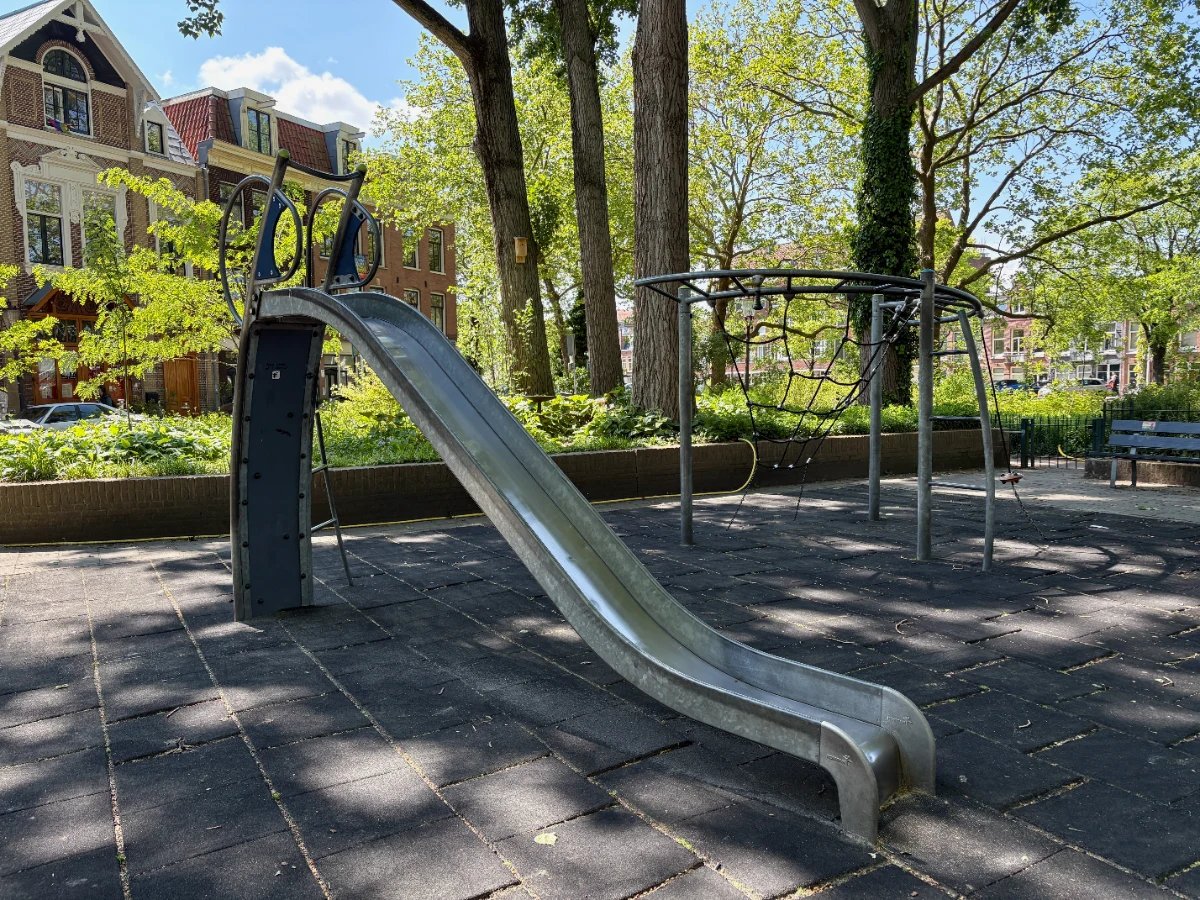 Shaded playground with a metal slide and rope climbing frame beneath large mature trees