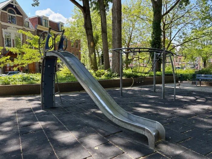 Shaded playground with a metal slide and rope climbing frame beneath large mature trees