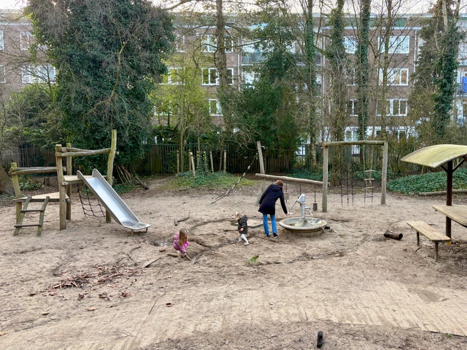 Mom and kids playing at nature playground with small wood and metal slides, water pump, picnic table