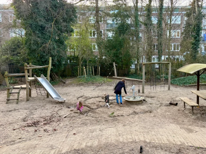 Mom and kids playing at nature playground with small wood and metal slides, water pump, picnic table