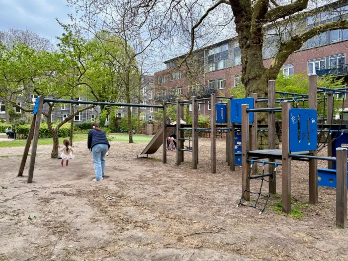 Mom pushes child on swing set attached to multi-level blue and wood play structure in urban park setting