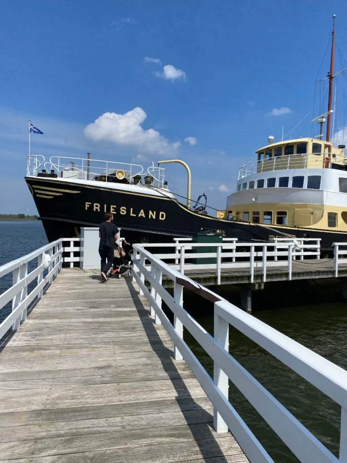 Man with stroller walks pier toward large black, yellow, and white steam boat with "Friesland" written on the side