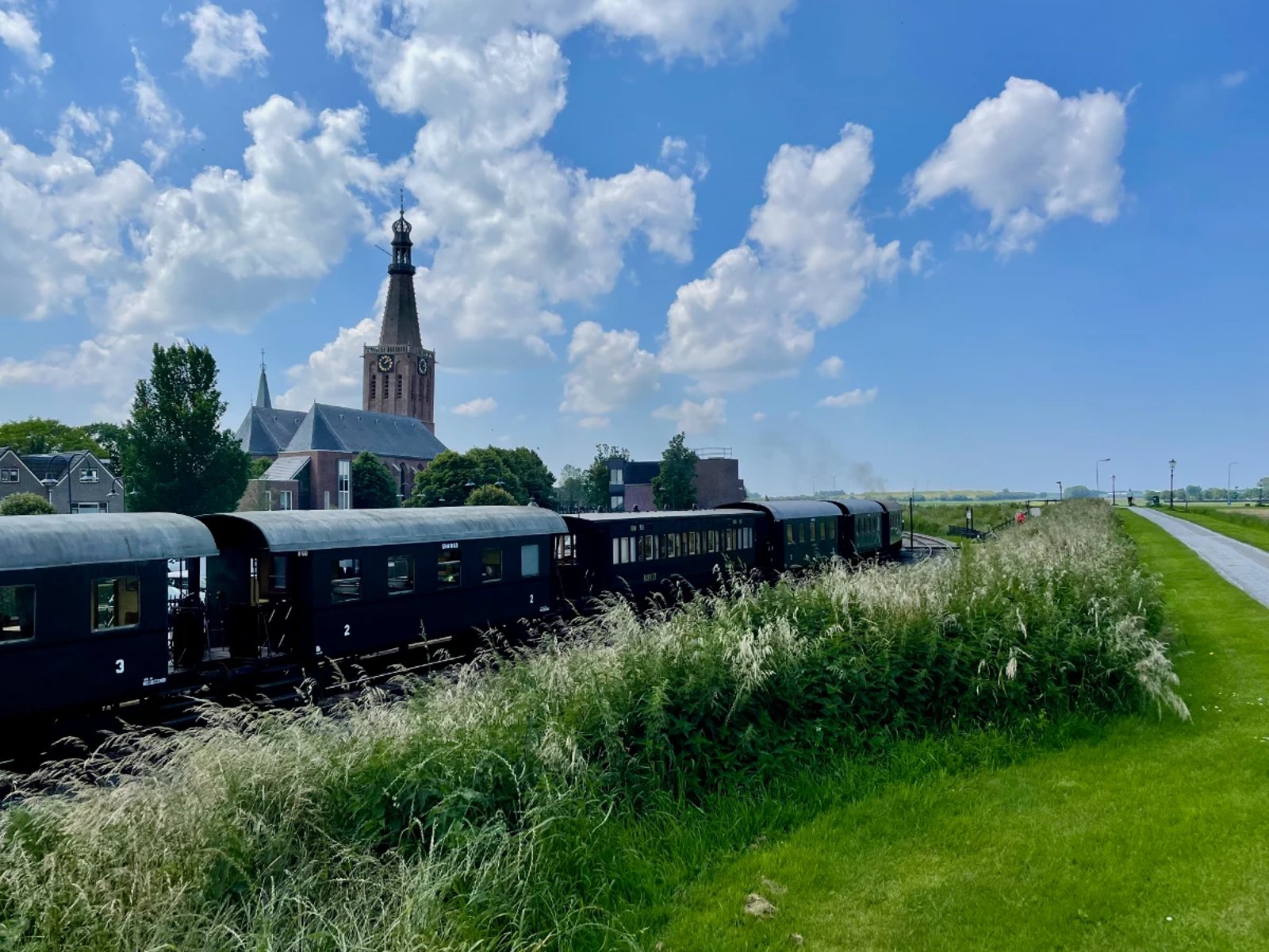 Historic steam train cars moving past a Dutch town with grass, bicycle path, and blue skies