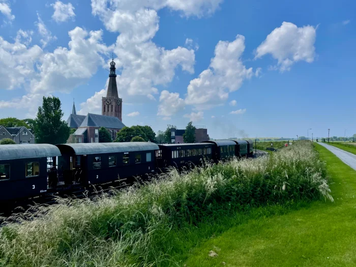 Historic steam train cars moving past a Dutch town with grass, bicycle path, and blue skies
