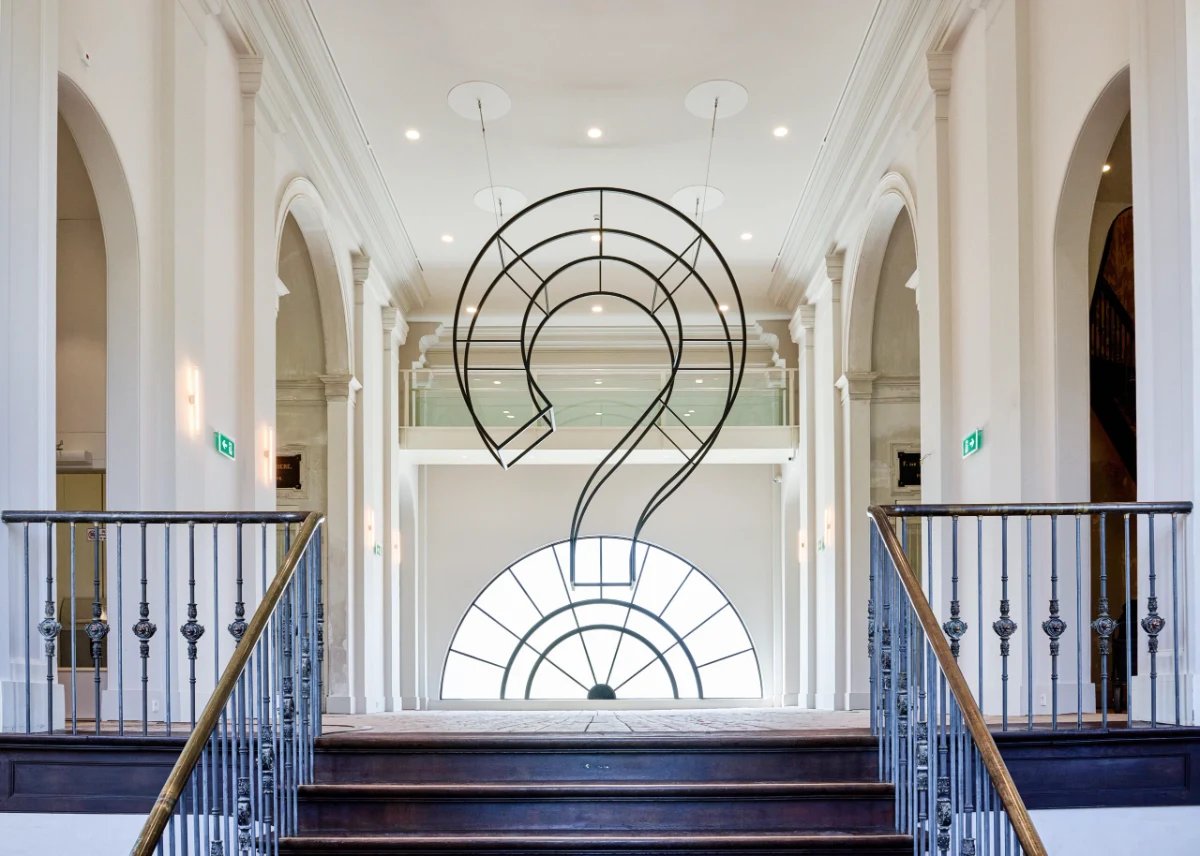 Large question mark sculpture made out of wire at the top of a staircase in a white room
