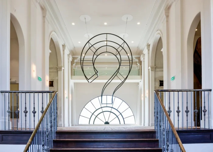 Large question mark sculpture made out of wire at the top of a staircase in a white room