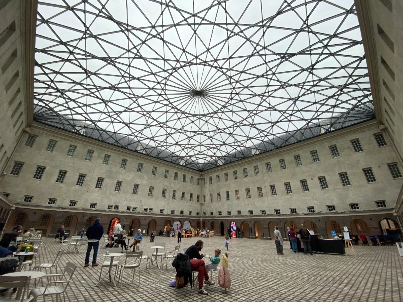 People sitting and dining in a large interior courtyard of a historic building that's covered with an intricate glass ceiling