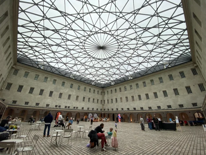 People sitting and dining in a large interior courtyard of a historic building that's covered with an intricate glass ceiling