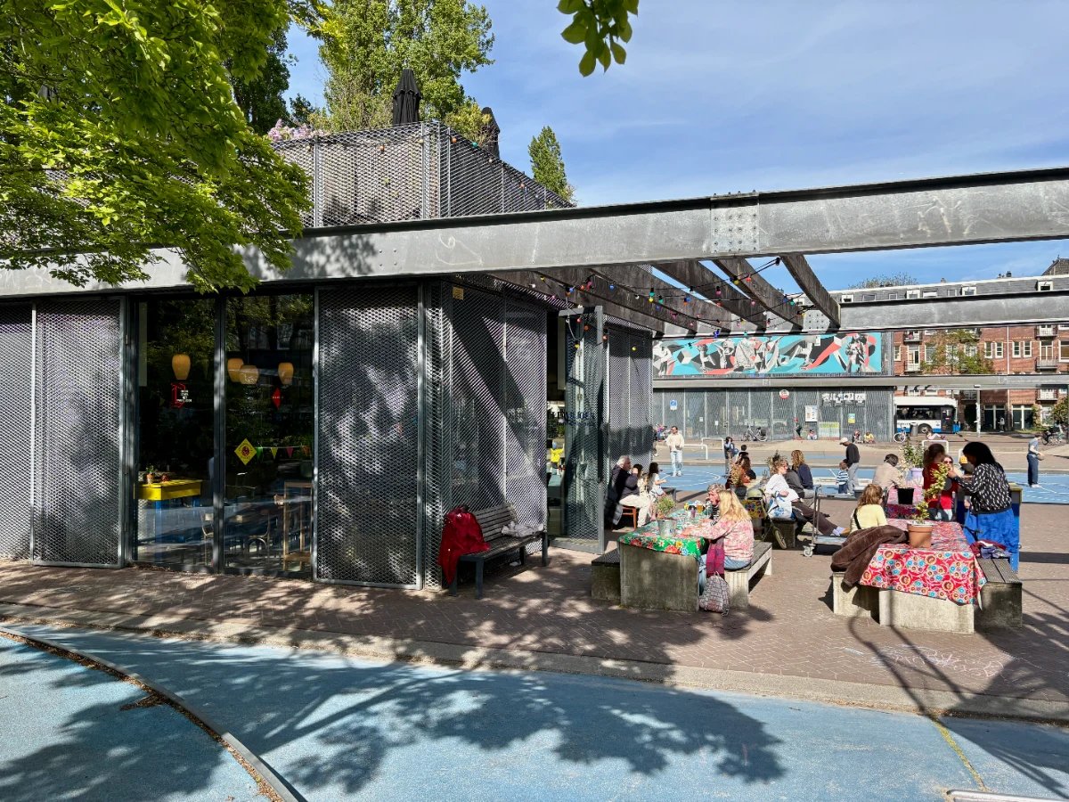 Modern metal cafe building with lots of people sitting at picnic tables outside beneath a steel pergola with playground in background