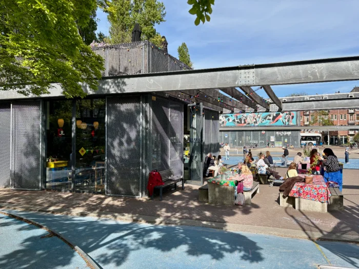 Modern metal cafe building with lots of people sitting at picnic tables outside beneath a steel pergola with playground in background