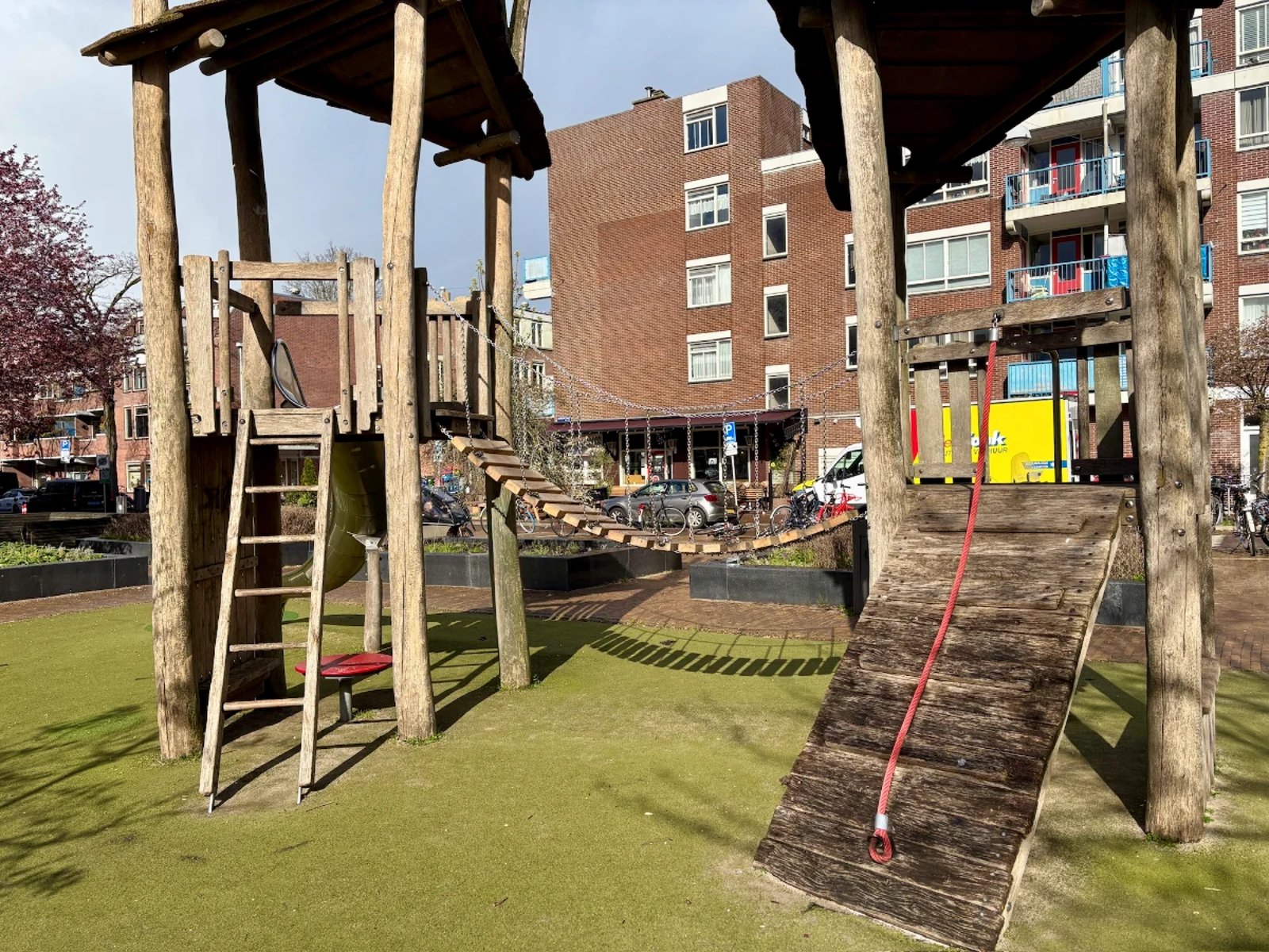 Wooden playground structure with two towers connected by a bridge and ladders and rope pull