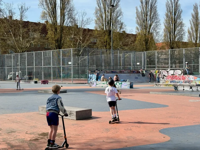 Kids on scooters at an urban skatepark surrounded by tall fence, trees, and residential buildings