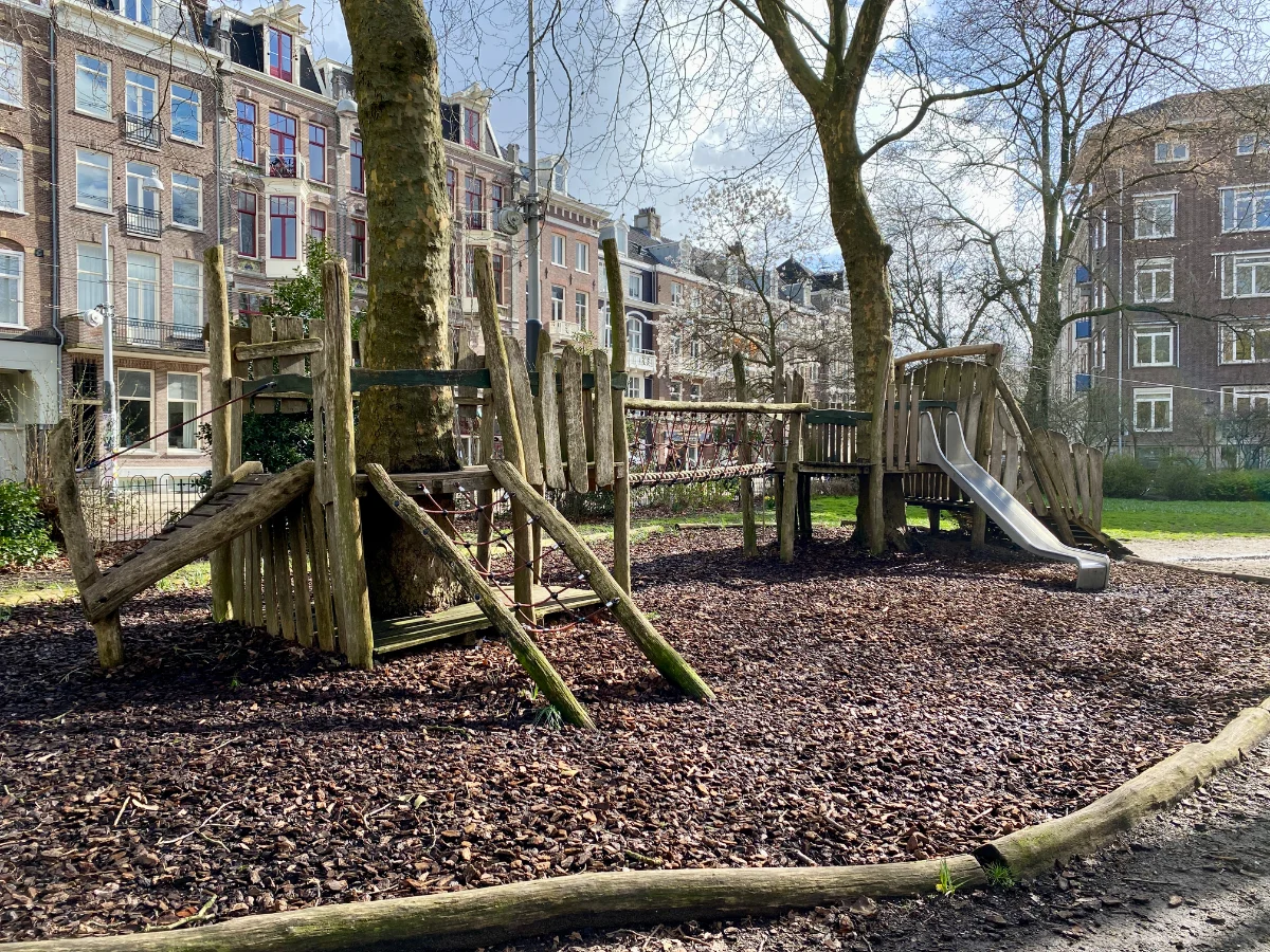 Wooden playground structure with ladders, bridges, and slides and mature trees around it