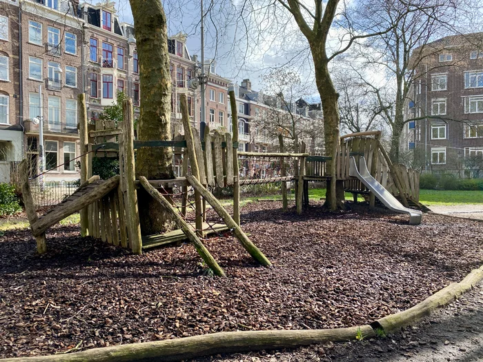 Wooden playground structure with ladders, bridges, and slides and mature trees around it