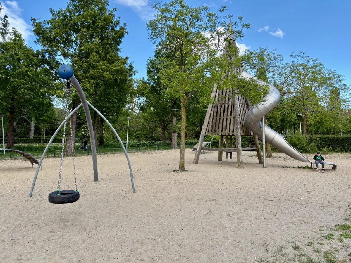 Tall wooden pyramid climbing tower with curved slide and tire swing in park setting with sand and trees around