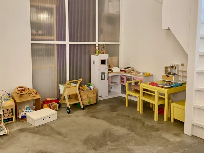 Play are with play kitchen, yellow table and chairs, shopping basket, steering wheel, and other toys in room with white walls and concrete floors