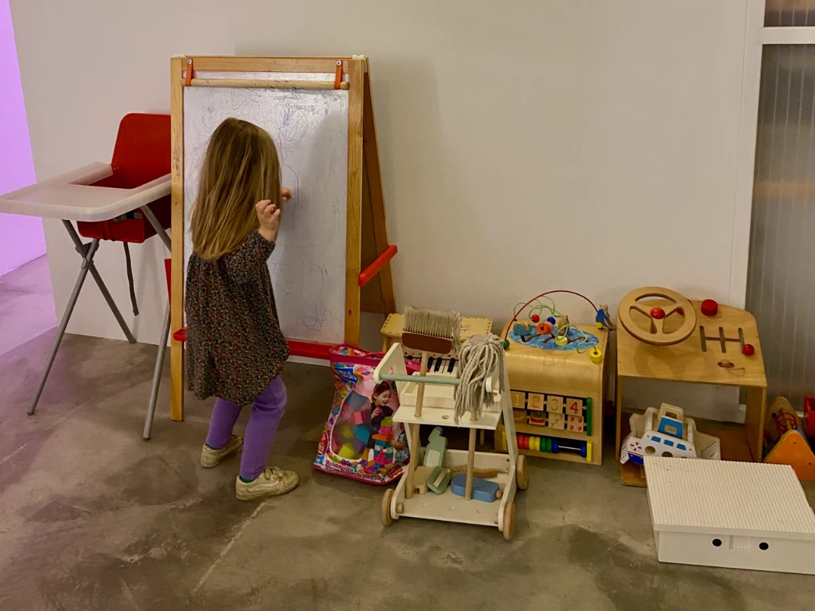 Girl coloring on a white board with high chair and other toys around