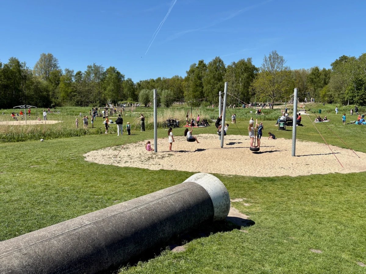 Kids playing on expansive playground with sandy area and a large concrete tunnel in the foreground