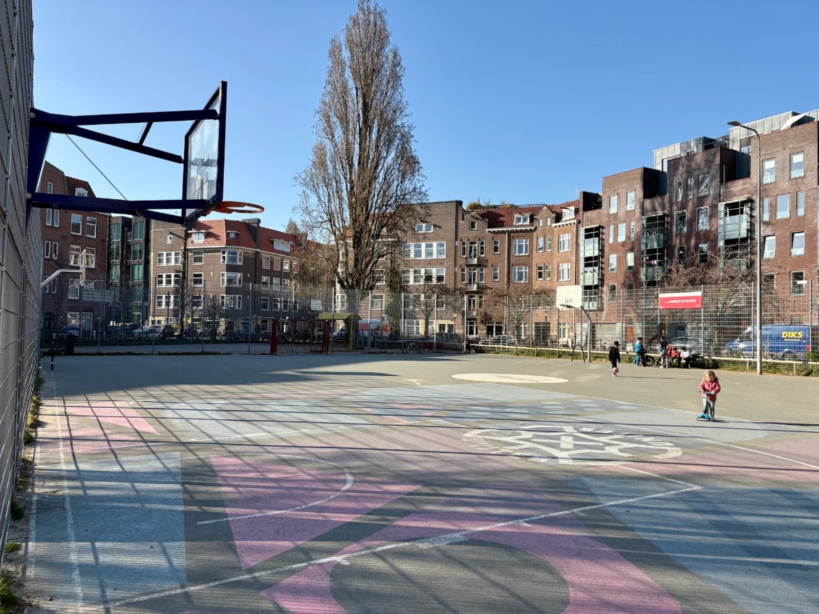 Toddler riding scooter on large open basketball court set in a residential plaza with red and blue shapes painted on the pavement