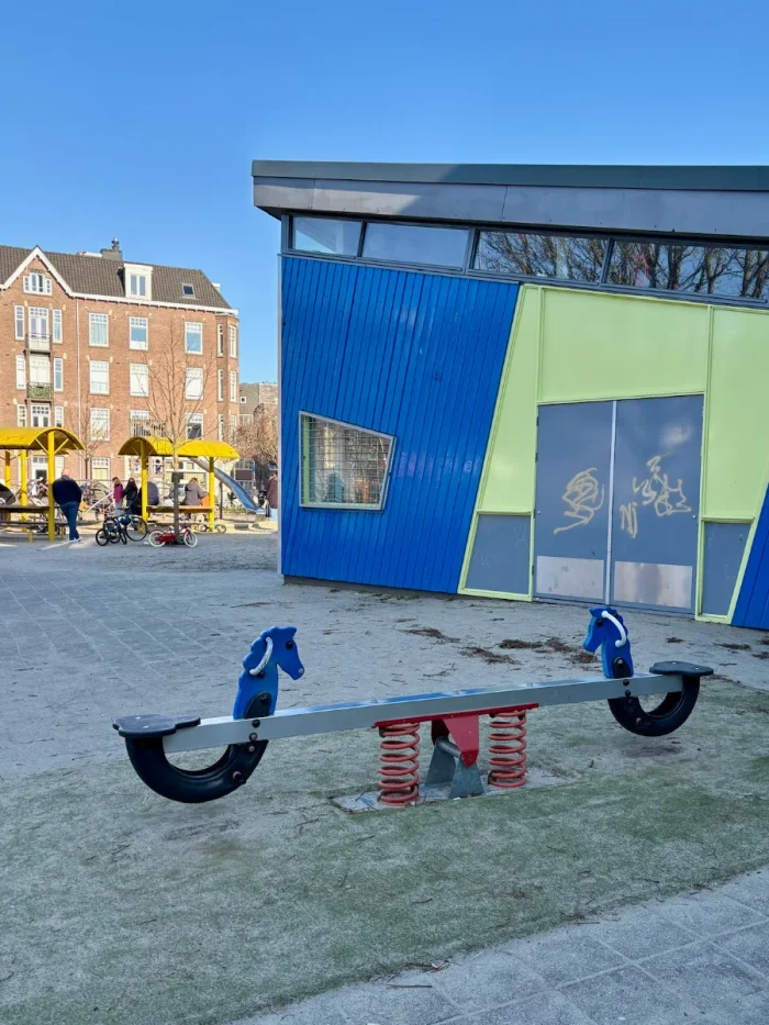 Blue horse seesaw with red springs in front of modern blue and yellow building