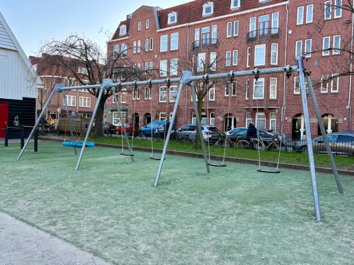 Large swing set with three swings and one nest swing with residential buildings behind