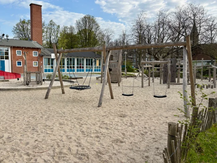Sandy playground with swings and nest swing and red brick and blue building behind