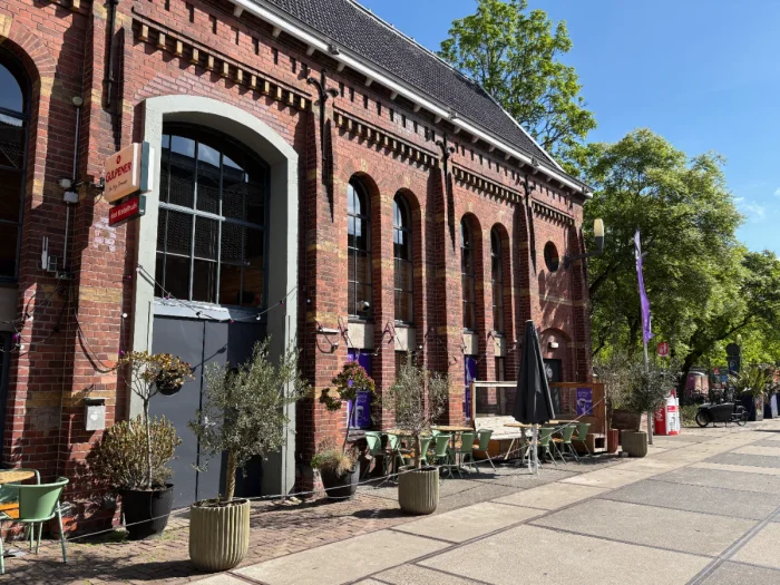 Industrial brick building on sunny day with tall, arched windows, outdoor seating, and potted plants